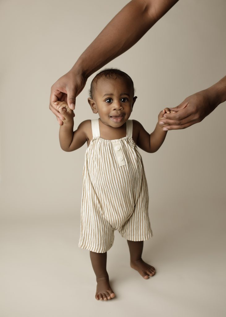9 month old baby standing with assistance by parent in photography studio